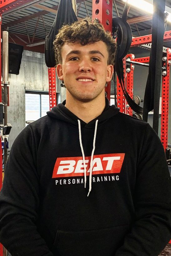 Young man at gym wearing Beat Personal Training hoodie, standing in front of fitness equipment.