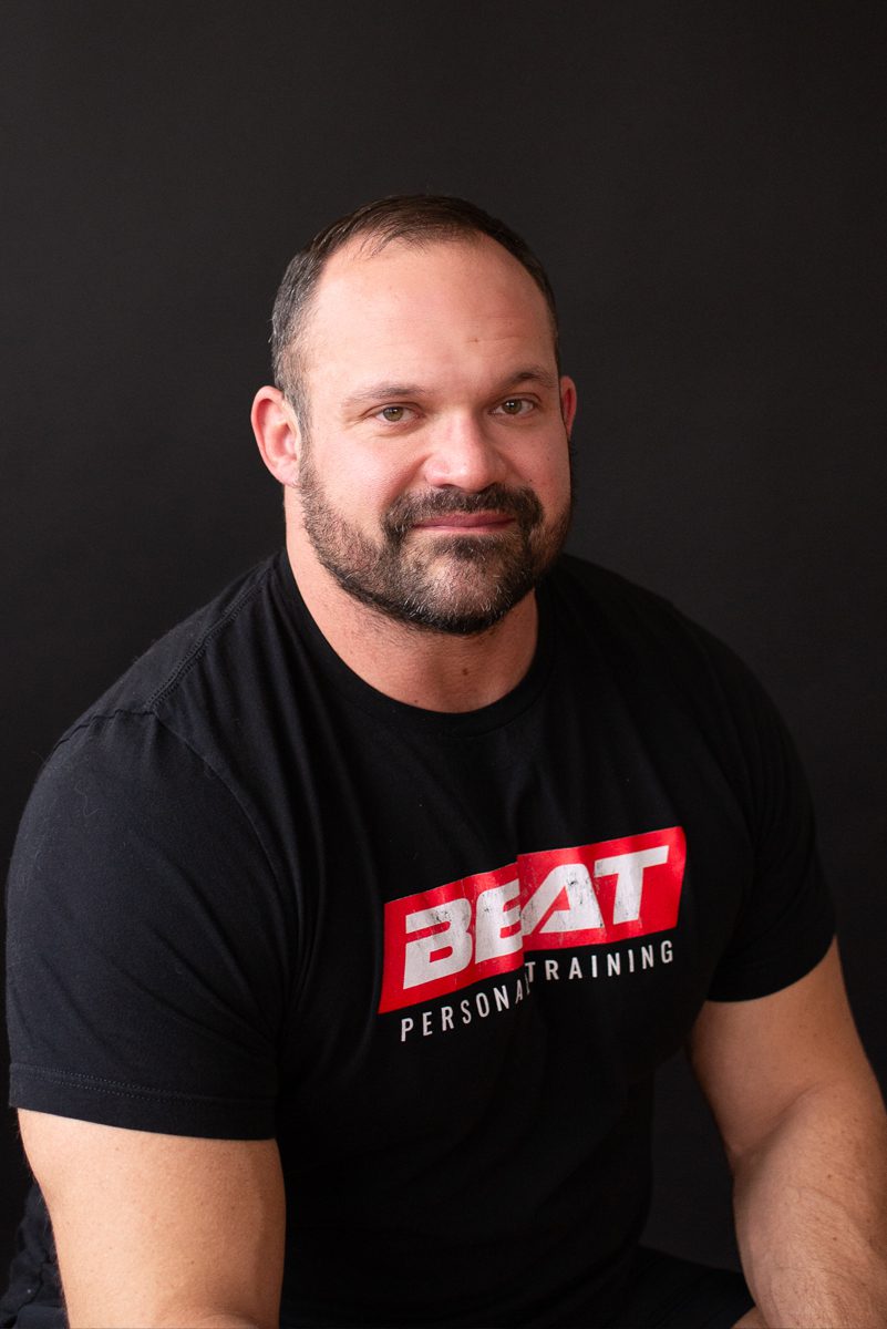 Man in a BEAT Personal Training shirt against a dark background.