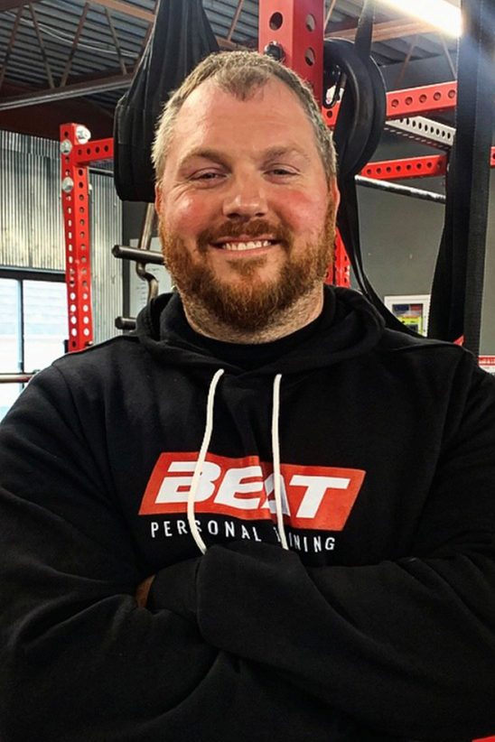 Man in gym with Beat Personal Training hoodie, smiling with arms crossed.