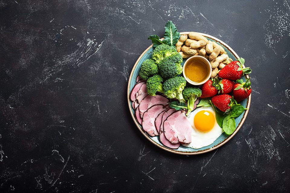 Healthy breakfast plate with ham, egg, broccoli, peanuts, and strawberries on dark background.
